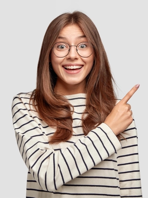 A joyful young woman wearing glasses and a striped sweater is pointing to her right with excitement.