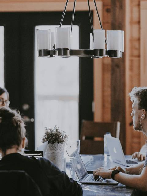A group of people working around a table in a cozy, well-lit room with large windows.