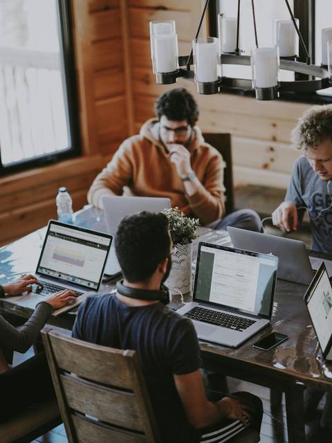 Three men focused on their laptops at a wooden table in a cozy, well-lit cabin.