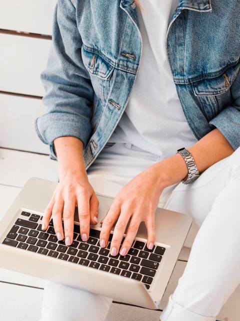 A person in a denim jacket and white pants typing on a laptop.