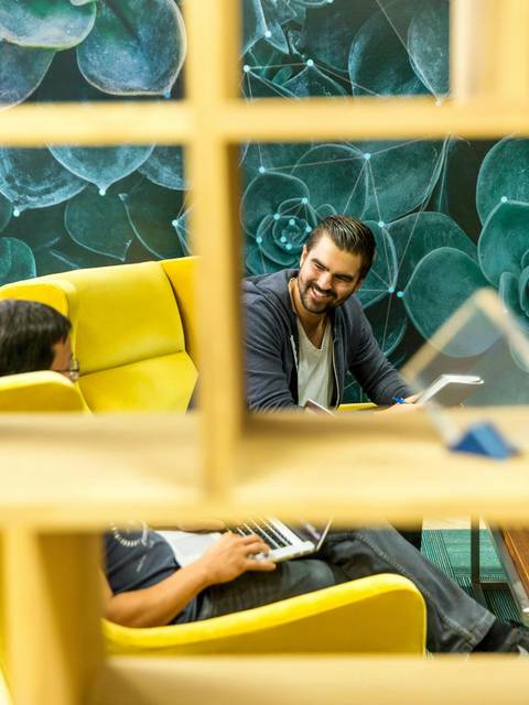 A man with a beard smiling and interacting with another person sitting on a yellow couch in a modern office space.