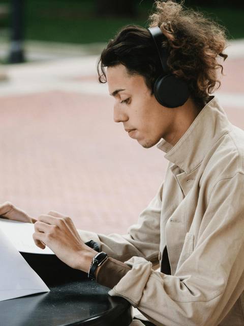 A young person is sitting at an outdoor table, wearing headphones and reading documents.