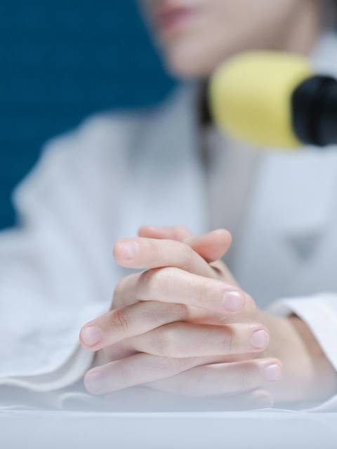 A person wearing a white jacket sitting at a desk with their hands clasped, speaking into a microphone with a yellow foam cover.