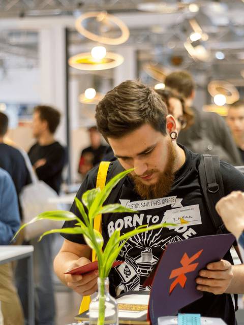 A young man reads a booklet at a busy convention center while holding a small plant.