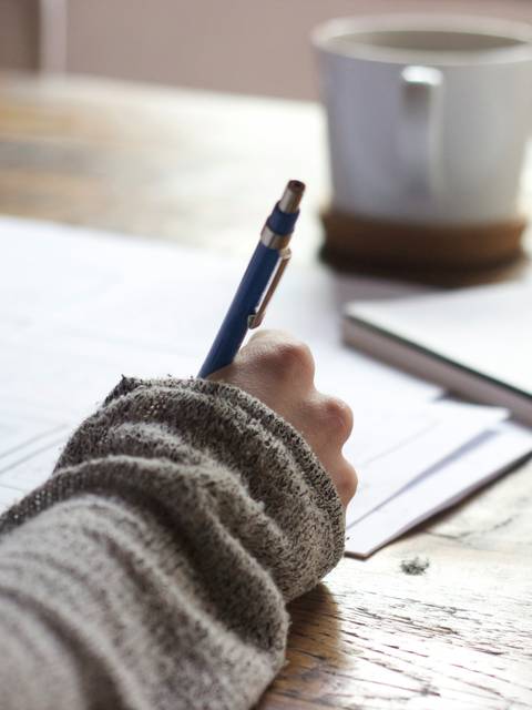 A person writing notes on a paper at a wooden table with a cup of coffee nearby.