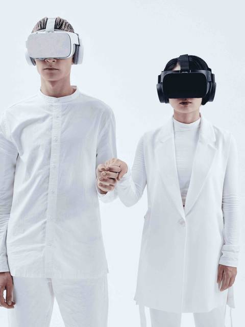 Two individuals wearing virtual reality headsets hold hands against a white background.