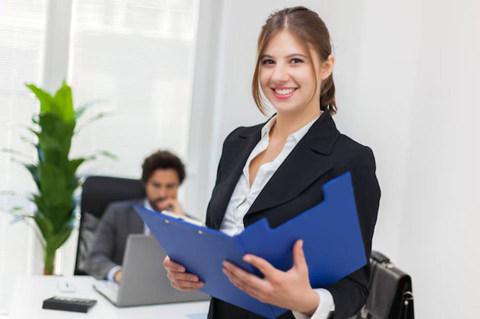 A smiling businesswoman holding a blue folder in an office environment.