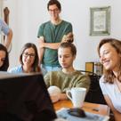 A group of young adults gathered around a laptop in a casual office setting, smiling and engaging in a discussion.