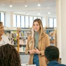 A young woman speaks while standing in a library with an older woman and several others listening.