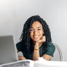 A smiling woman with curly hair sitting at a desk with a laptop and papers in a bright room.