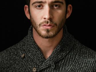 A portrait of a young man with a beard, wearing a dark knitted sweater against a black background.