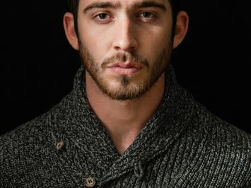 A portrait of a young man with a beard, wearing a dark knitted sweater against a black background.