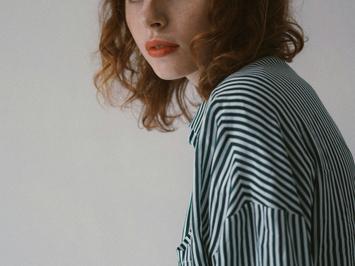 A portrait of a young woman with curly auburn hair, wearing a striped shirt, looking over her shoulder.
