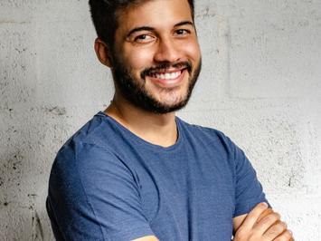 A young man with beard smiling confidently, standing against a white textured wall wearing a blue t-shirt with his arms crossed.