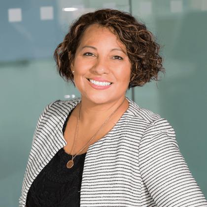 A portrait of a smiling woman with short curly hair, wearing a striped blazer and a black top, standing indoors.