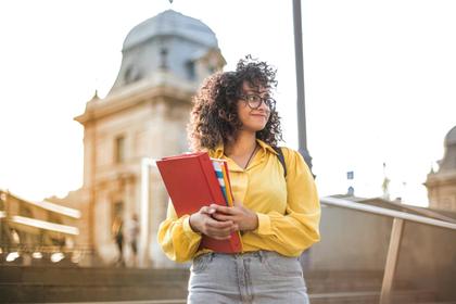 A young woman with curly hair, wearing glasses and a yellow hoodie, holds books while standing outdoors in a city.