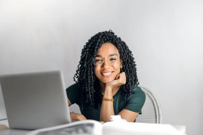 A smiling woman with curly hair sitting at a desk with a laptop and papers in a bright room.