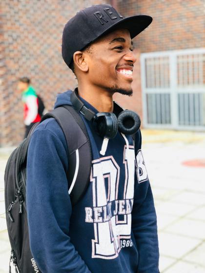 A young man with a cheerful expression wearing a baseball cap and headphones around his neck stands outdoors.
