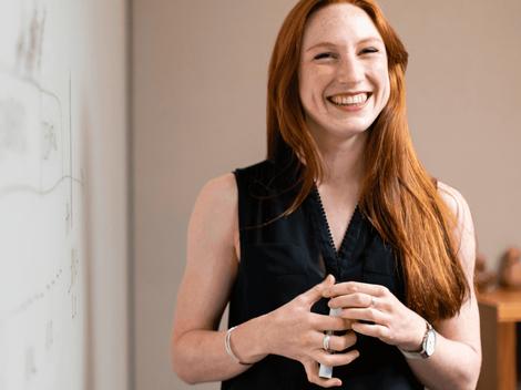 A joyful woman with red hair smiling in a room with a whiteboard.