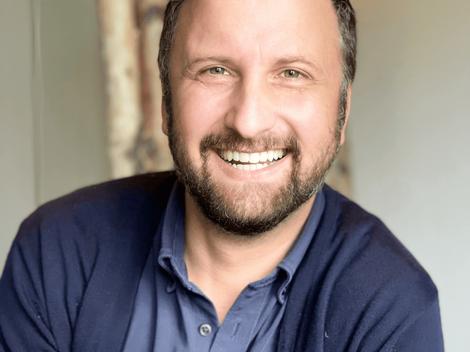 A smiling man with a beard wearing a dark blue shirt sitting in front of a softly blurred background.
