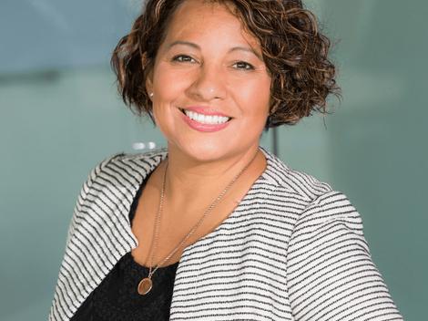 A professional portrait of a smiling woman with curly hair dressed in a striped jacket and a black top, taken indoors with a blurred background.