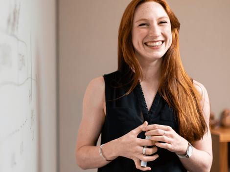 A smiling woman with red hair standing in front of a whiteboard in a meeting room.
