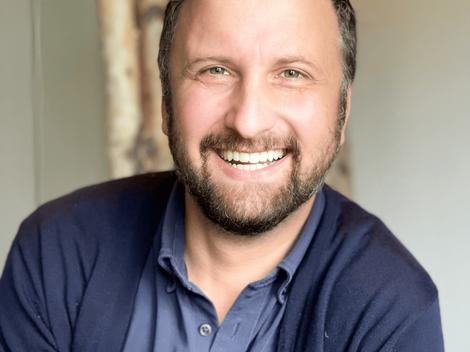 A portrait of a smiling man with a beard wearing a blue shirt.