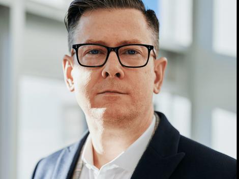 A professional portrait of a man with glasses wearing a blue blazer and white shirt, standing in an office environment.