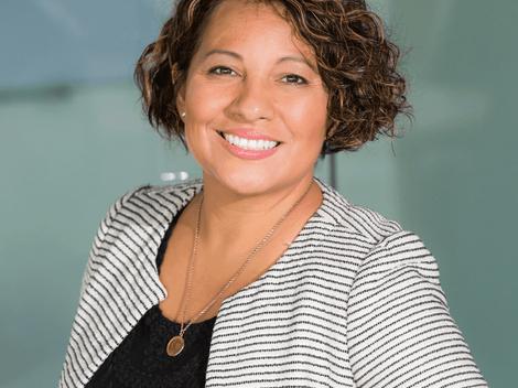 A portrait of a smiling woman with short curly hair, wearing a striped blazer and a black top, standing indoors.