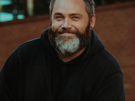 A portrait of a smiling man with a beard wearing a black hoodie.