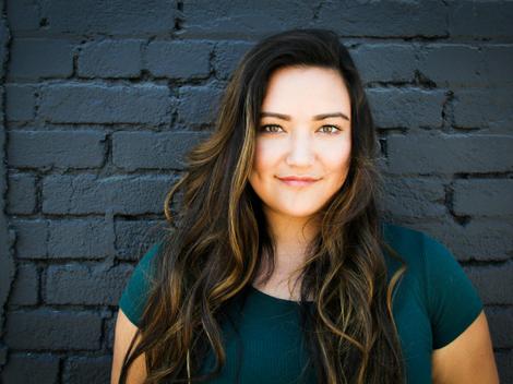 A woman with long hair smiling against a brick wall background.