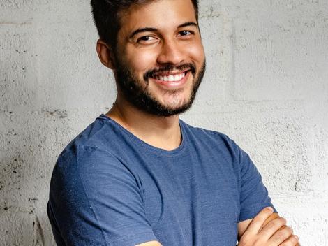 A smiling young man in a blue t-shirt standing with arms crossed against a textured white wall.