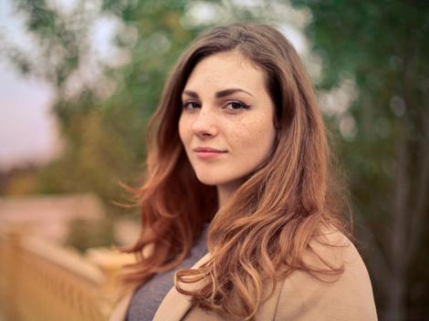 A portrait of a young woman with long brown hair, wearing a beige coat, standing outdoors with trees in the background.