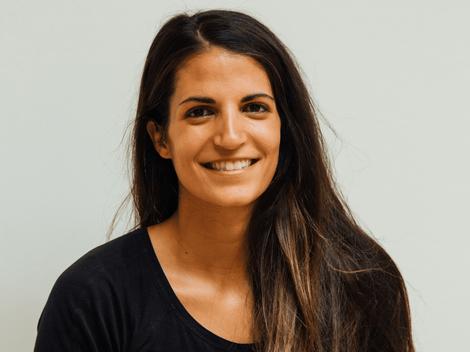 A smiling woman with long hair, wearing a black top, standing against a light-colored background.