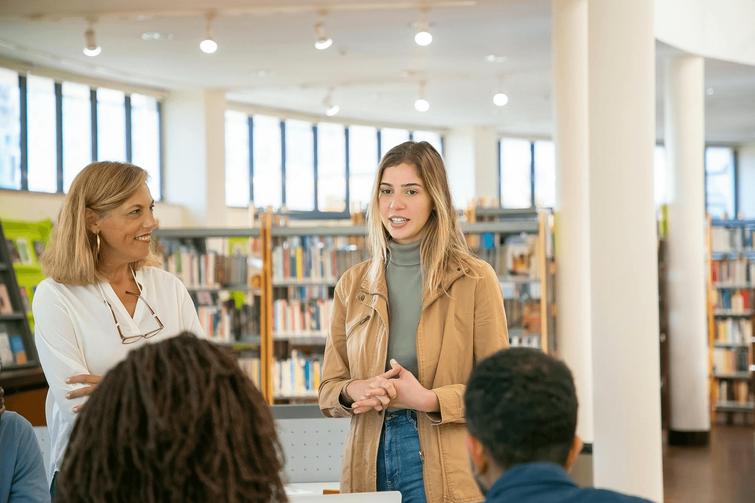 A young woman speaks while standing in a library with an older woman and several others listening.