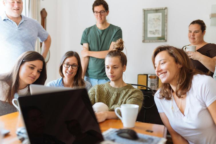 A group of young adults gathered around a laptop in a casual office setting, smiling and engaging in a discussion.