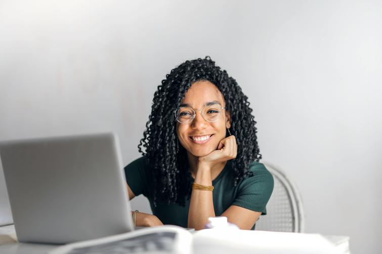 A smiling woman with curly hair sitting at a desk with a laptop and papers in a bright room.