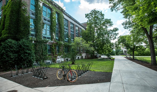 A peaceful outdoor scene at a university campus with a brick building, bike racks, and lush green trees.