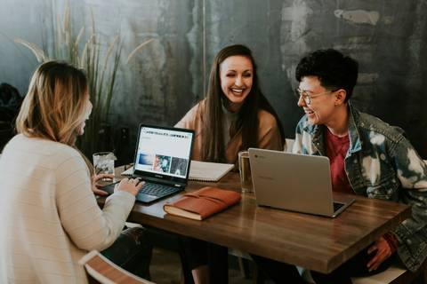 Three people smiling and engaging in a conversation at a table with laptops and a smartphone.