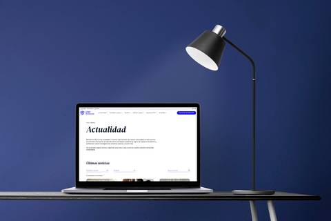 A laptop displaying a news website sits on a desk under a desk lamp against a blue background.